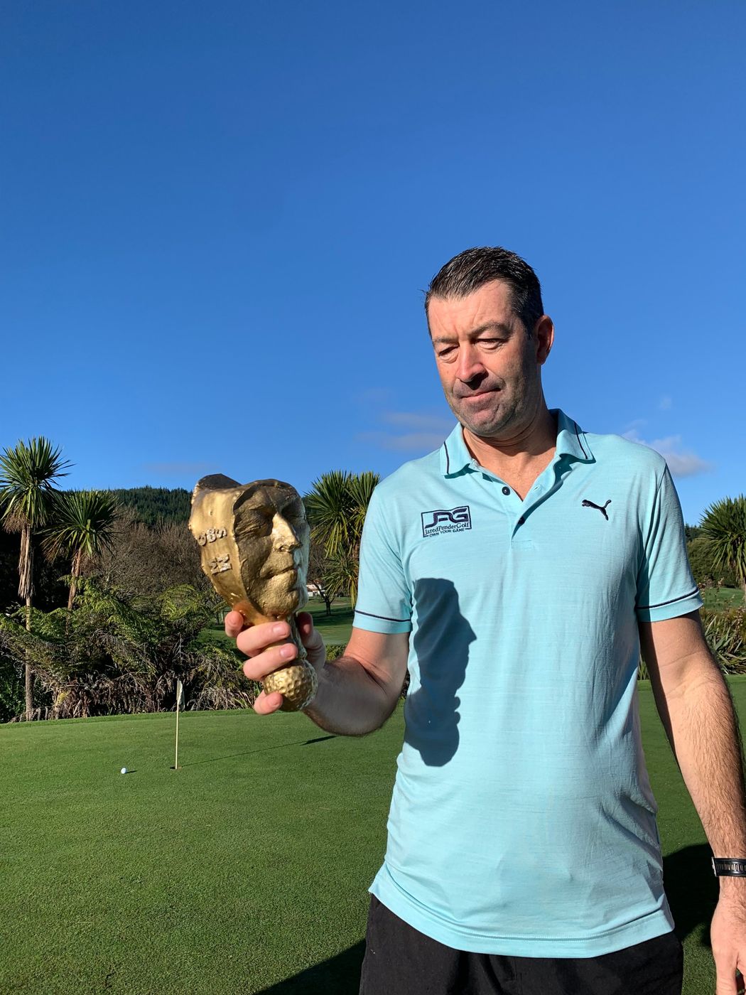 The Millar Moule Cup — a gilded face on a golf-ball base — held up on the green at Pauanui